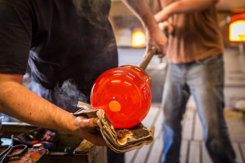 Glassmaker shaping molten glass in a furnace studio, is part of the handmade process behind an authentic Murano glass mirror.